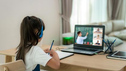 A grade school student at her home desk watches her teacher teach remotely on a laptop