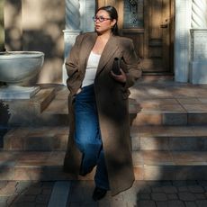 Marina wears a brown coat, white shirt, and jeans while posing with a handbag tucked under her arm