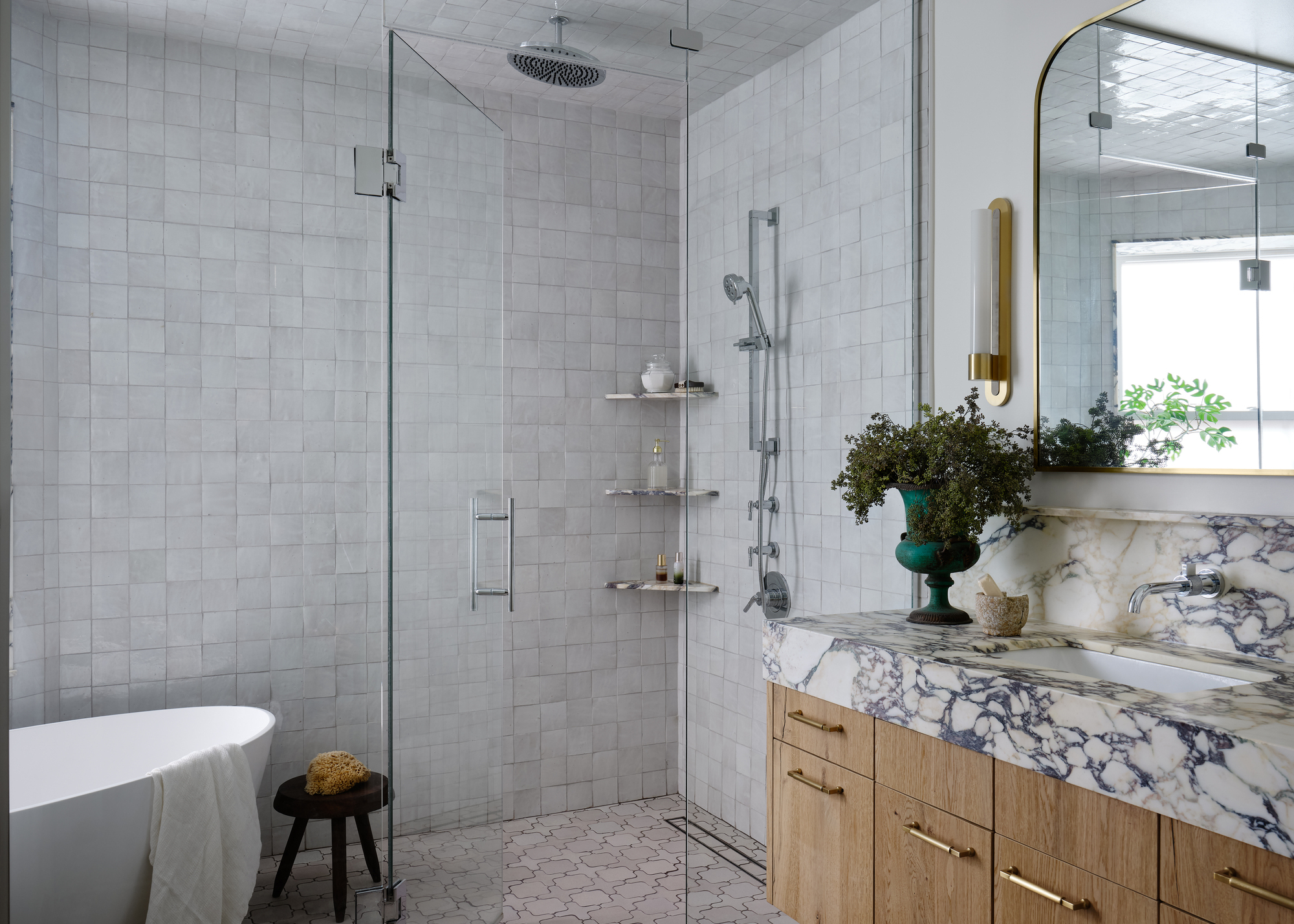 A walk-in shower in a glass enclosure fully tiled with while zellige tiles and a white bath in the corner