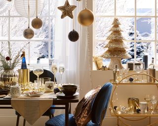 A gold and blue Art-Deco inspired festive dining area with straw Christmas tree in background above gold-framed bar cart