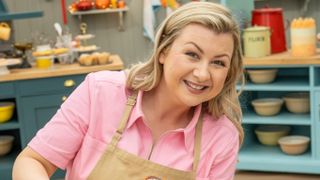 Rosie Ramsey smiling for the camera wearing a pink top and Bake Off apron