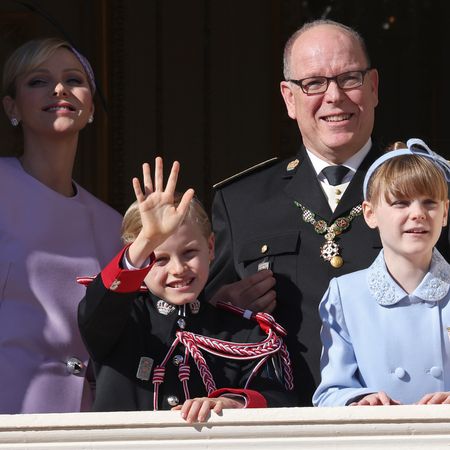 Princess Charlene, Prince Albert, Princes Jacques and Princess Gabriella wearing purple coats and military uniforms waving on a sunny balcony