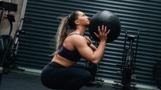 Aimee Cringle, the UK's fittest woman, crouches down and holds a medicine ball in her hands. She is in a dark gym; behind her we see metal shutter and airbikes.