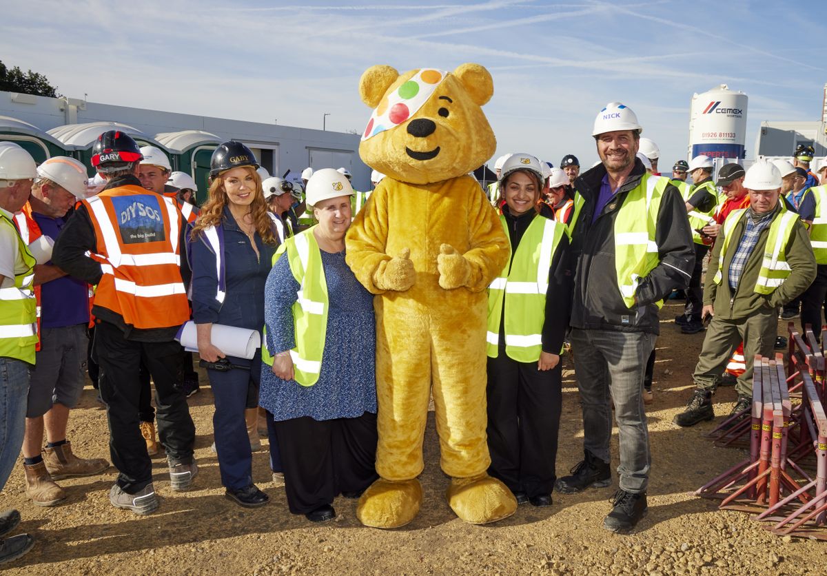 Pudsey Bear gives two thumbs up on the building site of this year&#039;s DIY SOS Children In Need special. Surrounding him, all in high-vis jackets and hard hats, are designer Gaby Blackman, Flavia and Sharan from the charity Getaway Girls, and presenter Nick Knowles