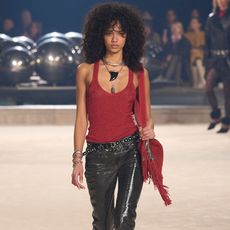 Isabel Marant runway model wearing red tank top, leather pants, studded belt, and fringe bag