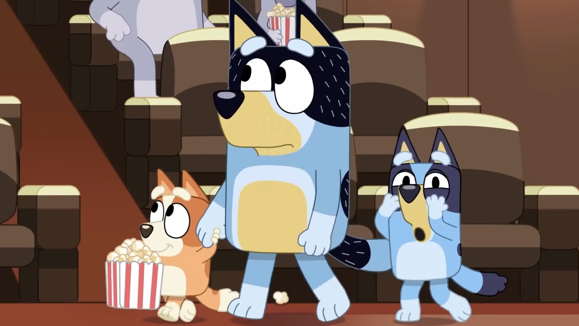 Bandit walking into theater screening room with Bingo holding popcorn and Bluey with hands over her eyes