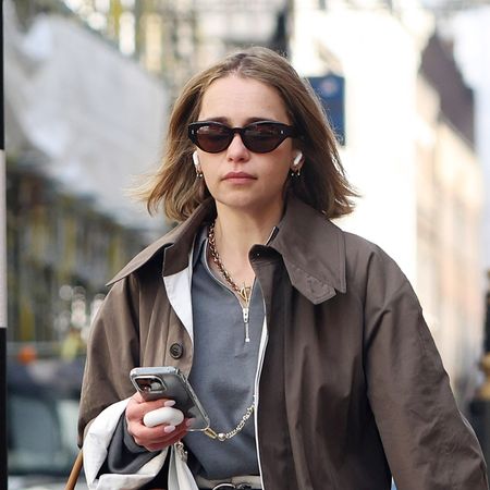 Emilia Clarke wearing a tan trench coat, gray half-zip pullover, white jeans, and tan Prada belt bag 