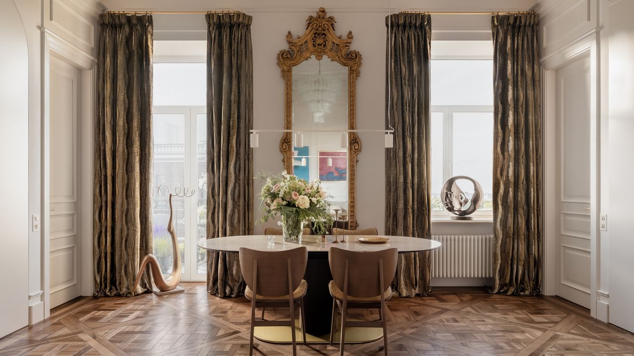 Image of white dining room with high ceiling and crown molding and hardwood floors. There is a small oval dining table and floor-to-ceiling golden curtains.