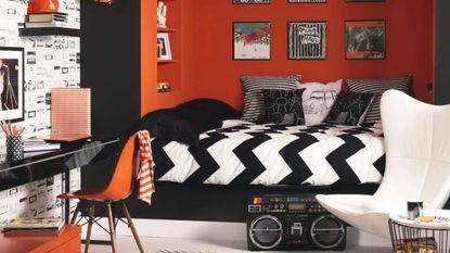 Retro black and orange bedroom, painted black and orange walls, bed built into alcove raised on platform, framed wall mounted album covers, white wing backed chair large ghetto blaster, black gloss desk, wallpaper with cassette tape print, white glass pendant light