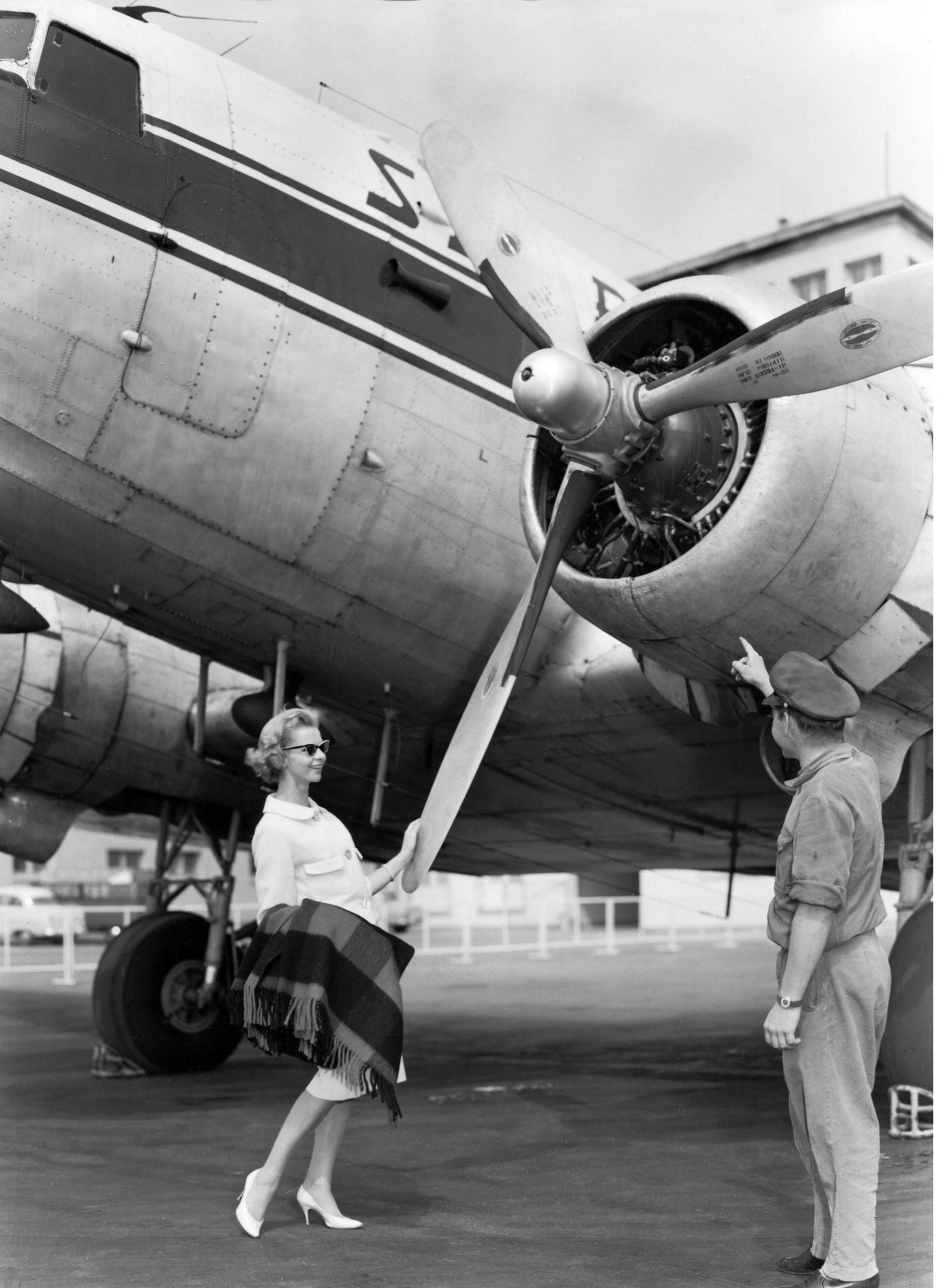A woman and a pilot standing in front of a vintage airplane. She is in sunglasses and touching the propellor. He is in a pilot's hat and facing away from the camera and pointing upwards
