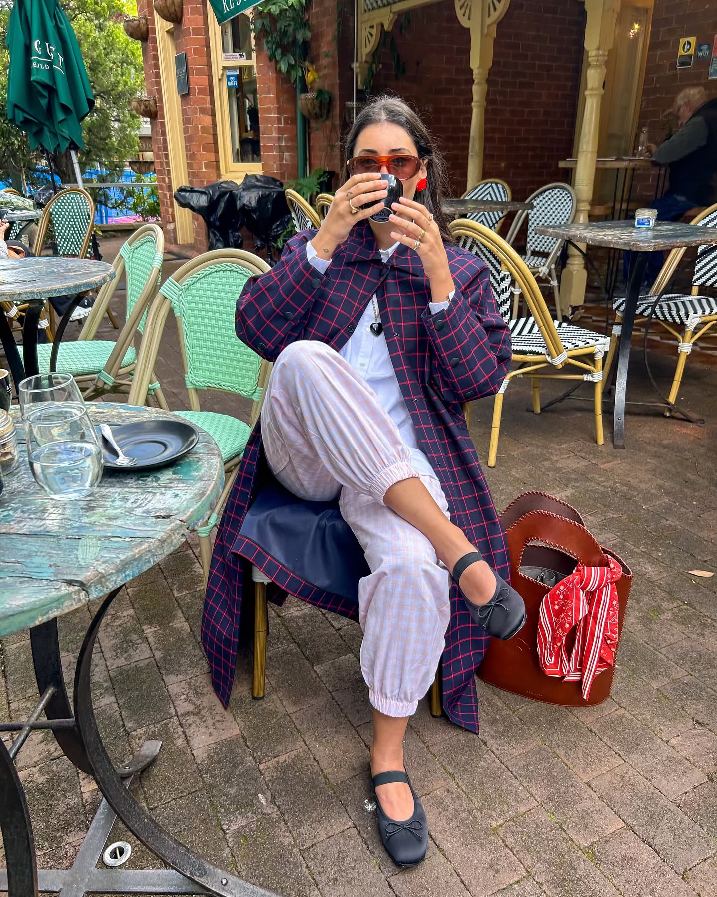 Influencer sits outside drinking a coffee wearing pink gingham trousers with black Mary Janes and a checked jacket.