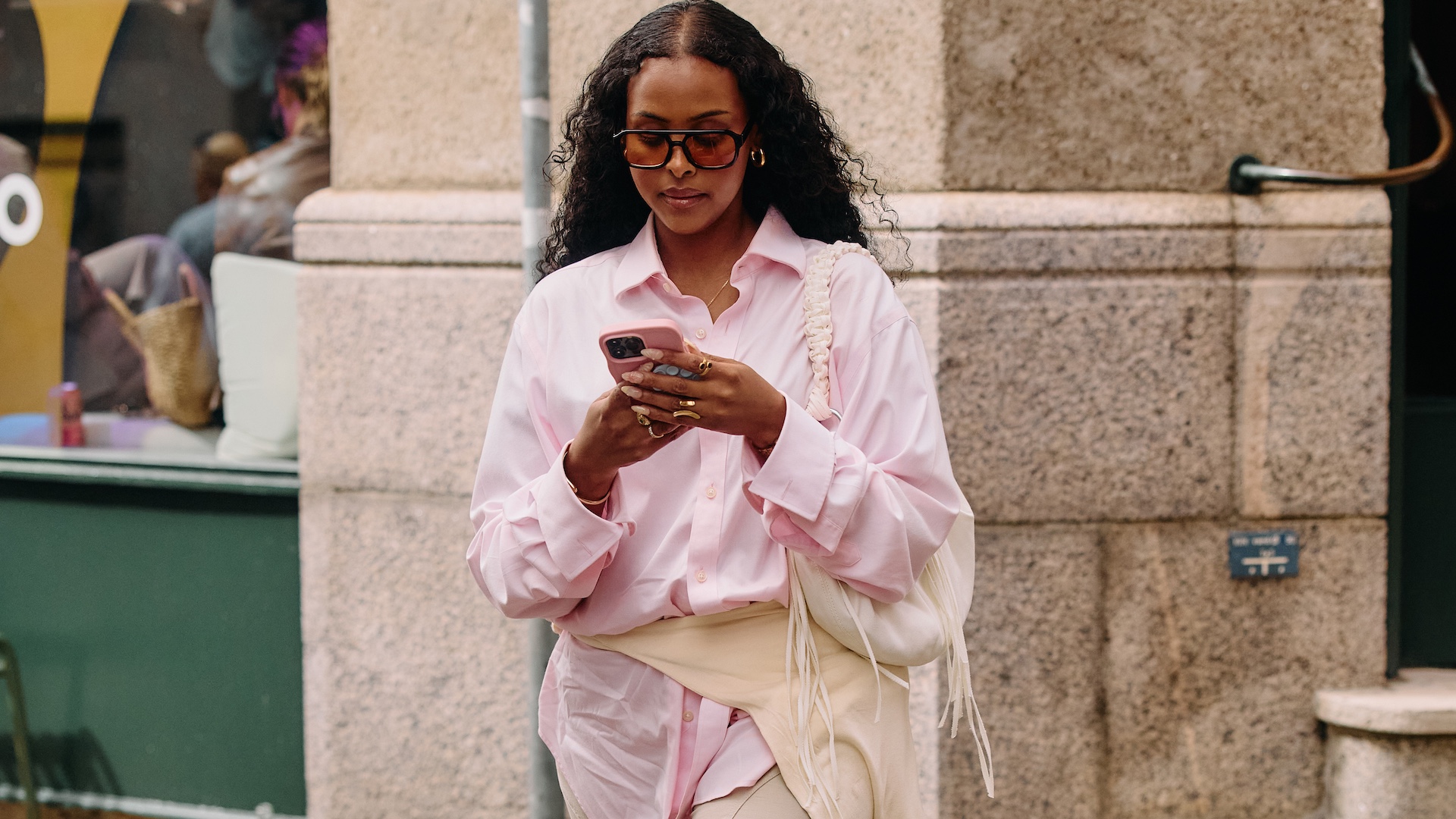 street style shot of a woman looking at her phone wearing aviator sunglasses, a pink button-up shirt, cream shoulder bag, and a beige sweater tied around her waist