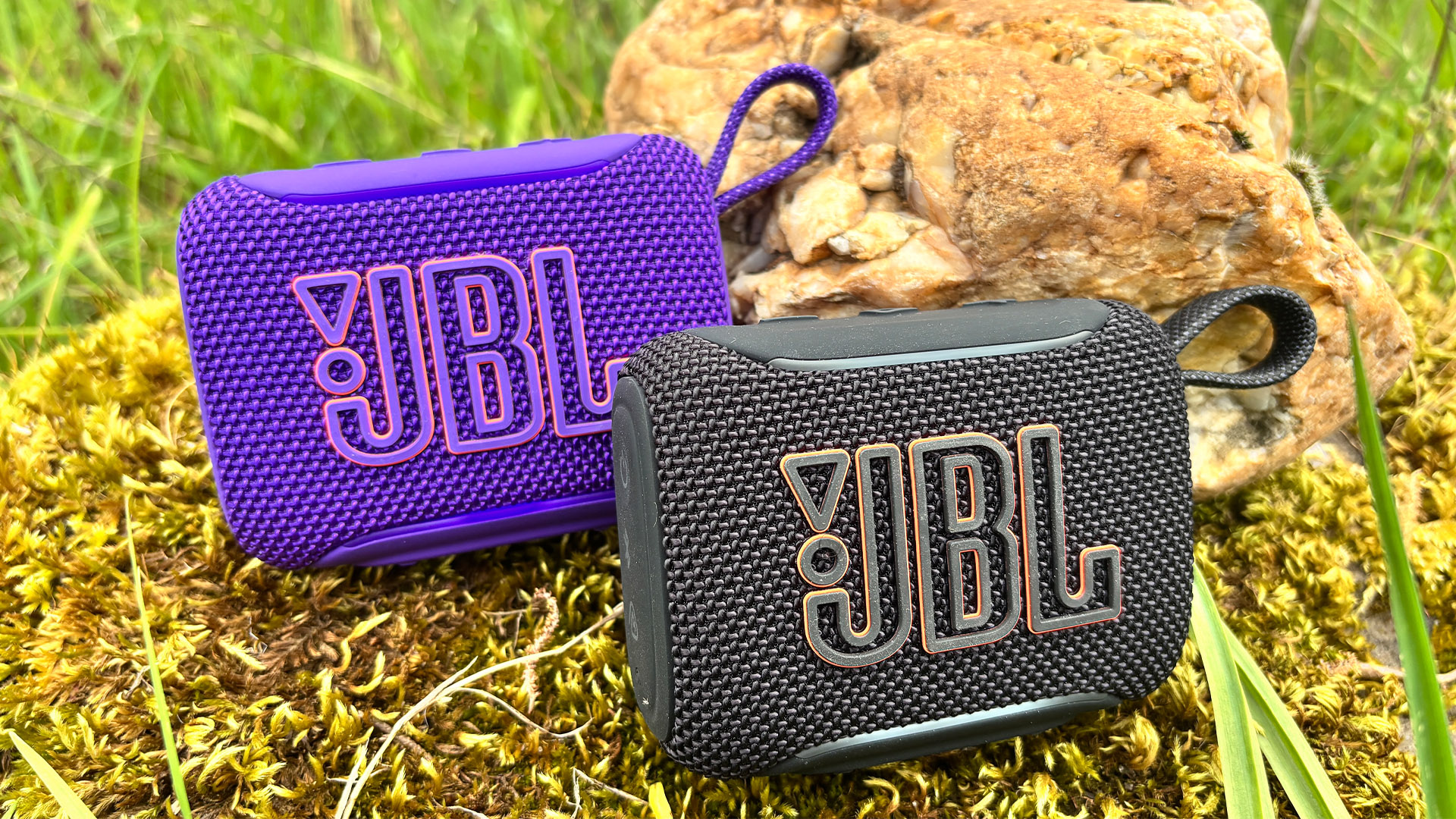 Two JBL Go 5 speakers, one in purple finish behind the one in black finish on mossy stone