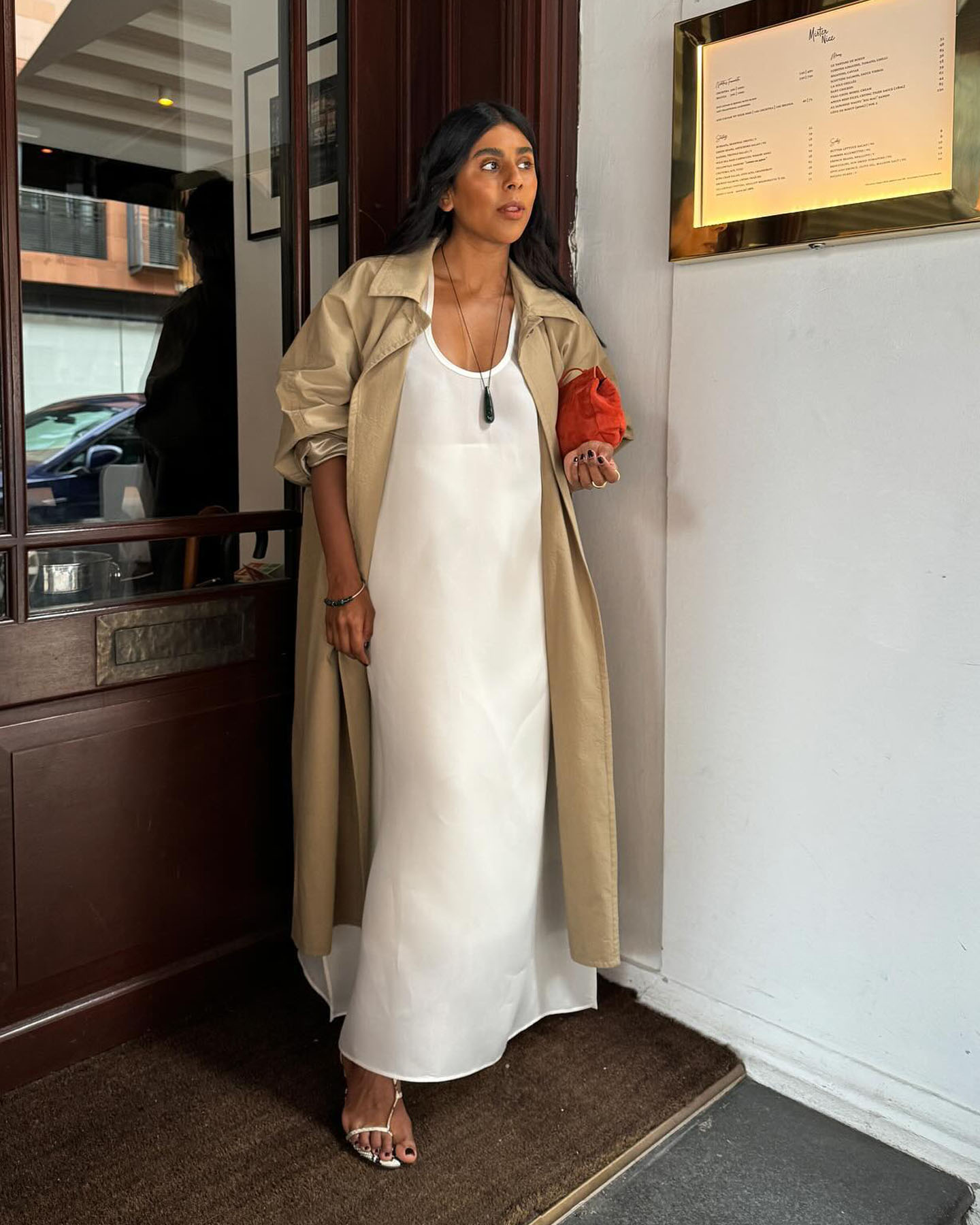 British female influencer Monikh Dale poses in the doorway of a London restaurant wearing a maxi trench coat, red suede clutch bag, white maxi dress, and white strappy sandals.