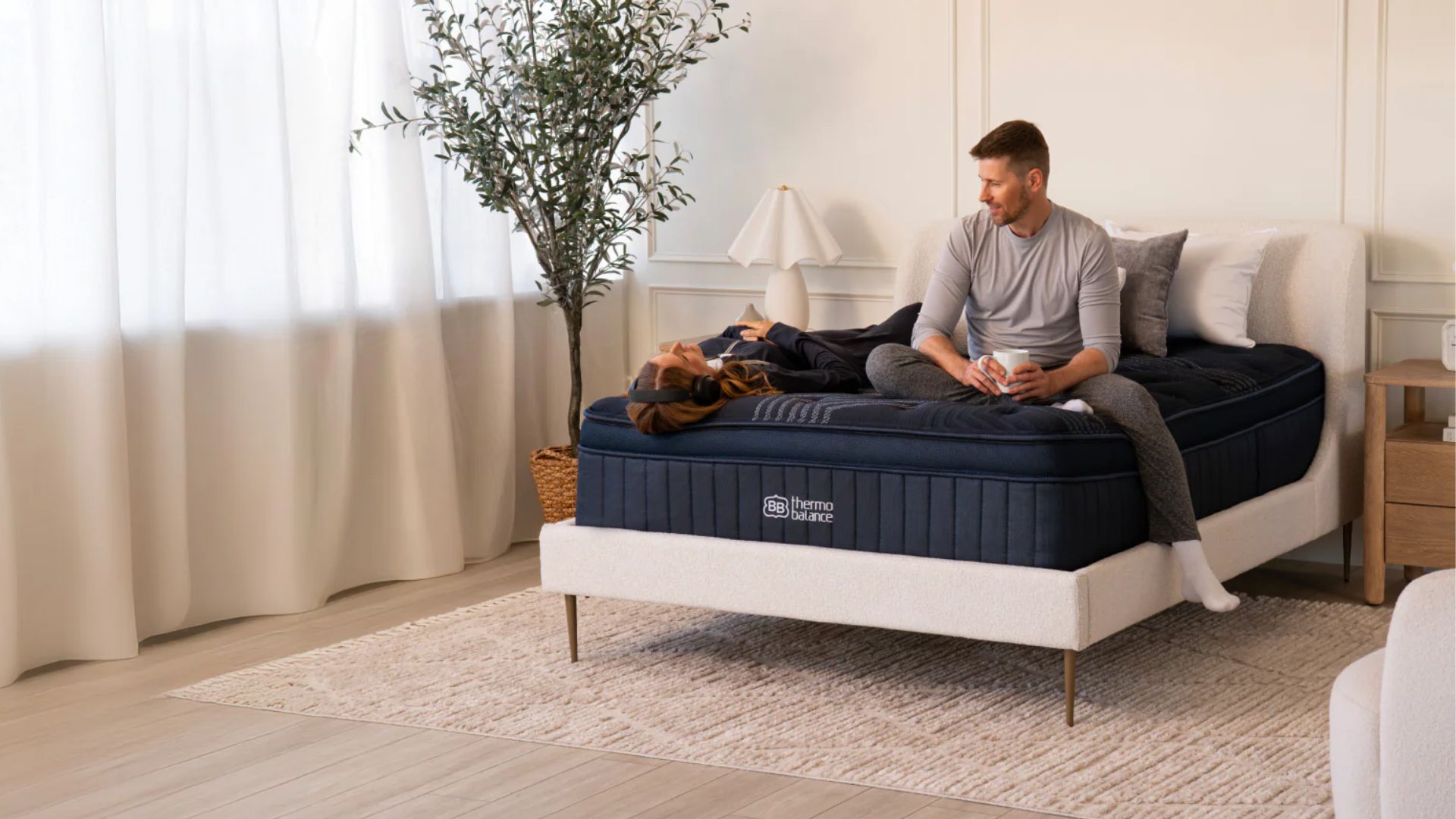 A man sits up on the ThermoBalance mattress while holding a mug, and a woman with headphones lies in the supine positions next to him.