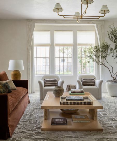 A light and airy living room with white walls, large windows, a rust-colored sofa and a large wood coffee table with books and decor. A textured rug and two cream armchairs. 