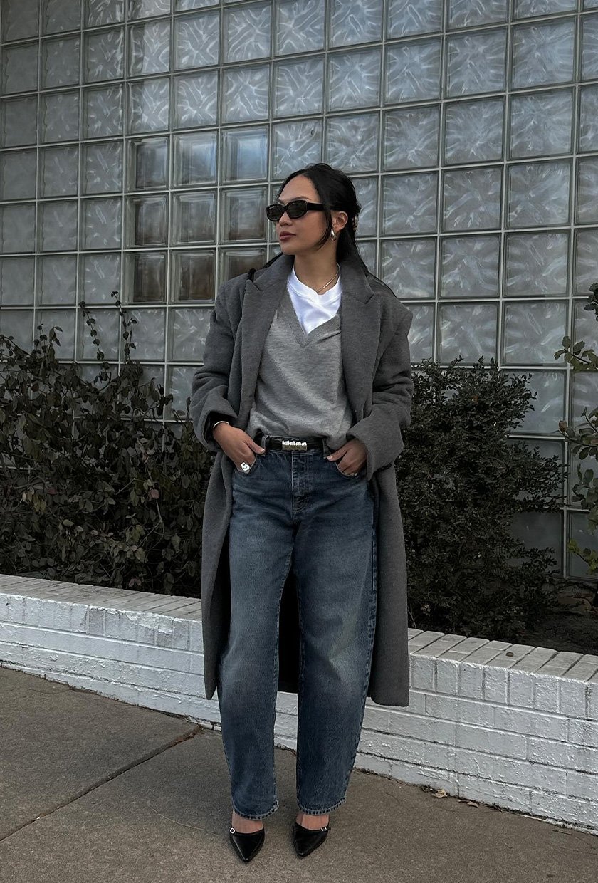 A woman standing on the sidewalk wearing a long gray wool coat over a gray V-neck with a white T-shirt underneath, a black belt, distressed barrel-leg jeans, and black pointed pumps.