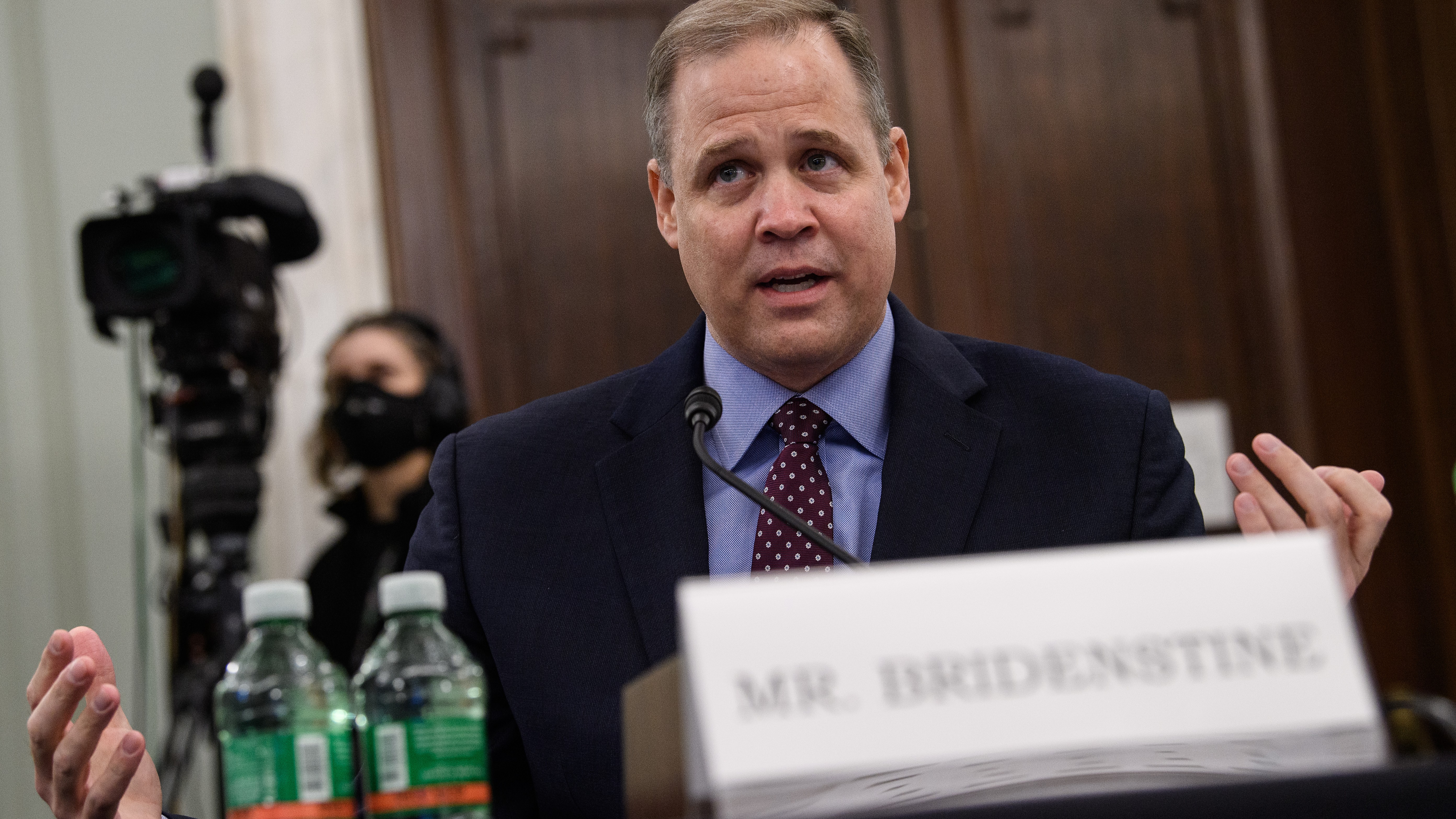 A man in a suit sits behind a table with open arms.