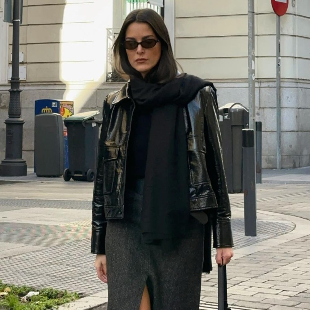 Influencer @leiasfez wears a patent leather jacket with a wool midi pencil skirt and black knee-high leather boots. She accessorises with sunglasses, a dark scarf and a light beige bag.