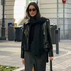 Influencer @leiasfez wears a patent leather jacket with a wool midi pencil skirt and black knee-high leather boots. She accessorises with sunglasses, a dark scarf and a light beige bag.
