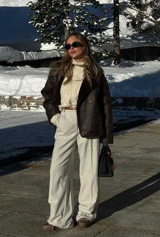 what to wear in 30-degree temperatures, shown in a photo of a woman standing outside in the snow wearing a shearling coat over a chunky white cable knit sweater, brown belt, white trousers, and brown snow boots