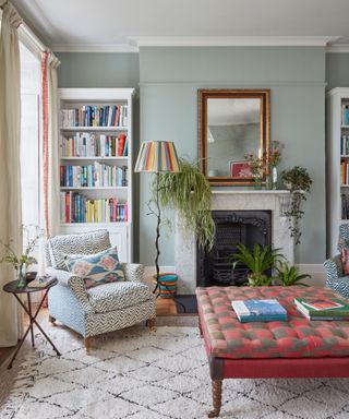 Gray-green living room, white fireplace, mirror on mantel, pink and gray patterned ottoman, bookshelf, cozy rug and armchair