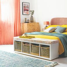 A white bedroom with a pink bed and an IKEA KALLAX storage unit used as a bench with storage baskets placed at the foot of the bed