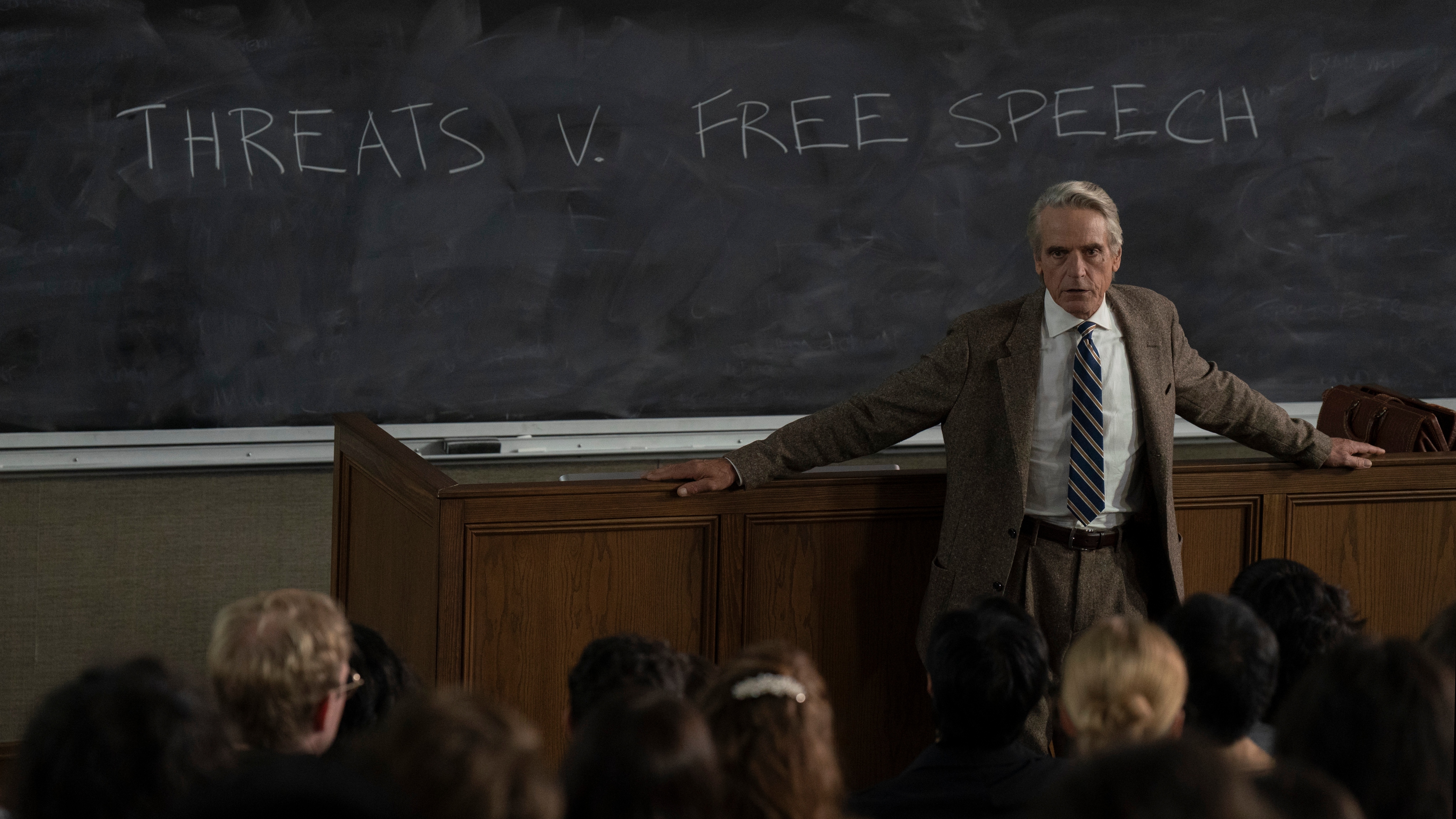 Jeremy Irons as Alex's father in The Morning Show season 4 - standing in front of a blackboard in a lecture hall with the words 'threats v free speech' in chalk behind him