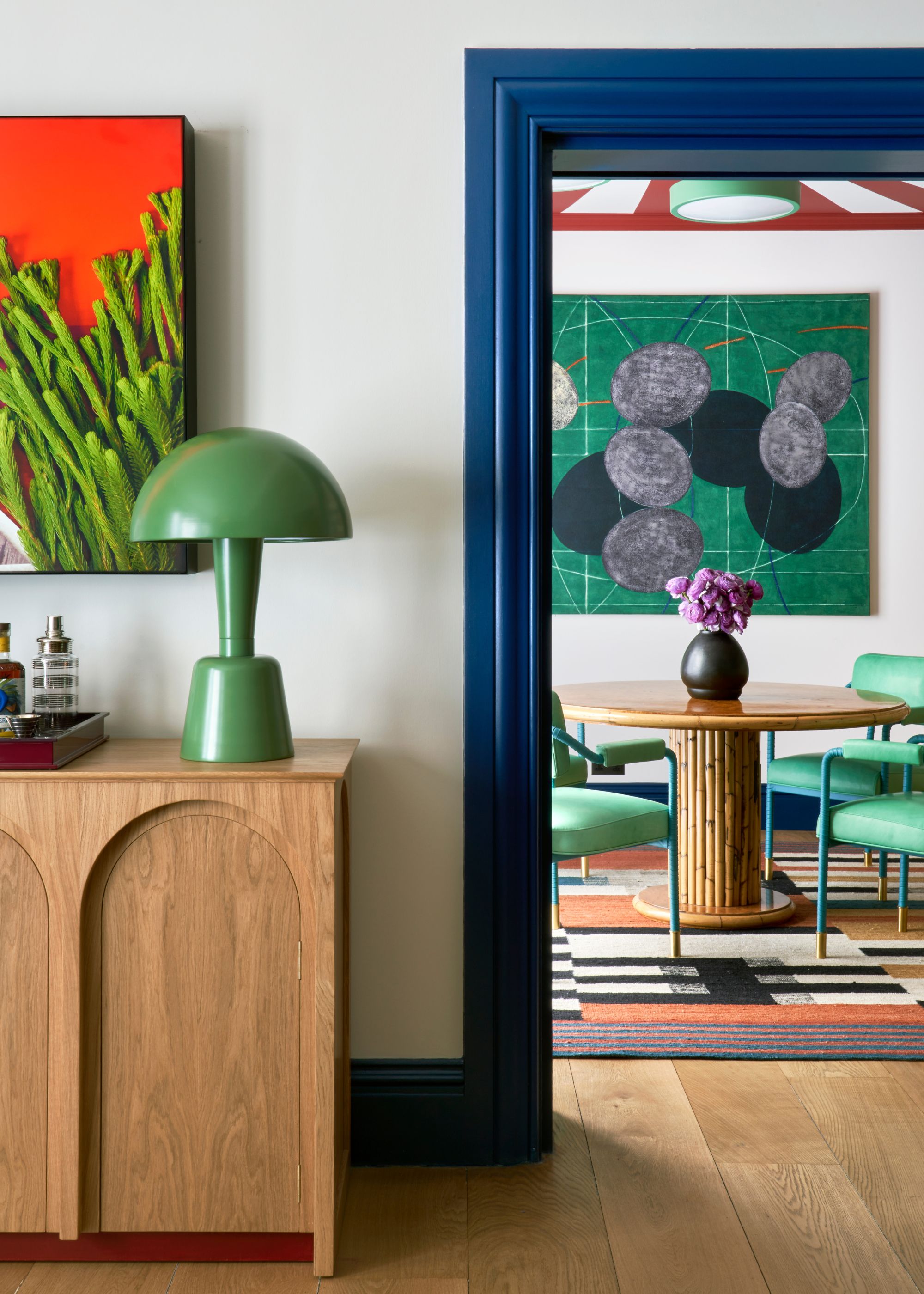 A doorway leading into a dining room with white walls, dark blue trim, wooden flooring, and a wood sideboard with a green modern lamp and a red and green artwork above it. A large green artwork in the dining room with a round wood table, green chairs, and a colorful rug.