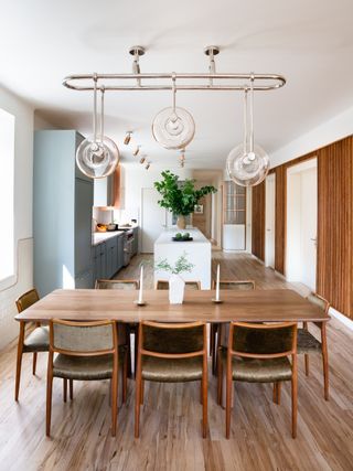 An open plan dining and kitchen area with a wooden table and chairs, glass pendants above, a white kitchen island, shelves, drawers, oven, stove, a potted plant and wooden floors.