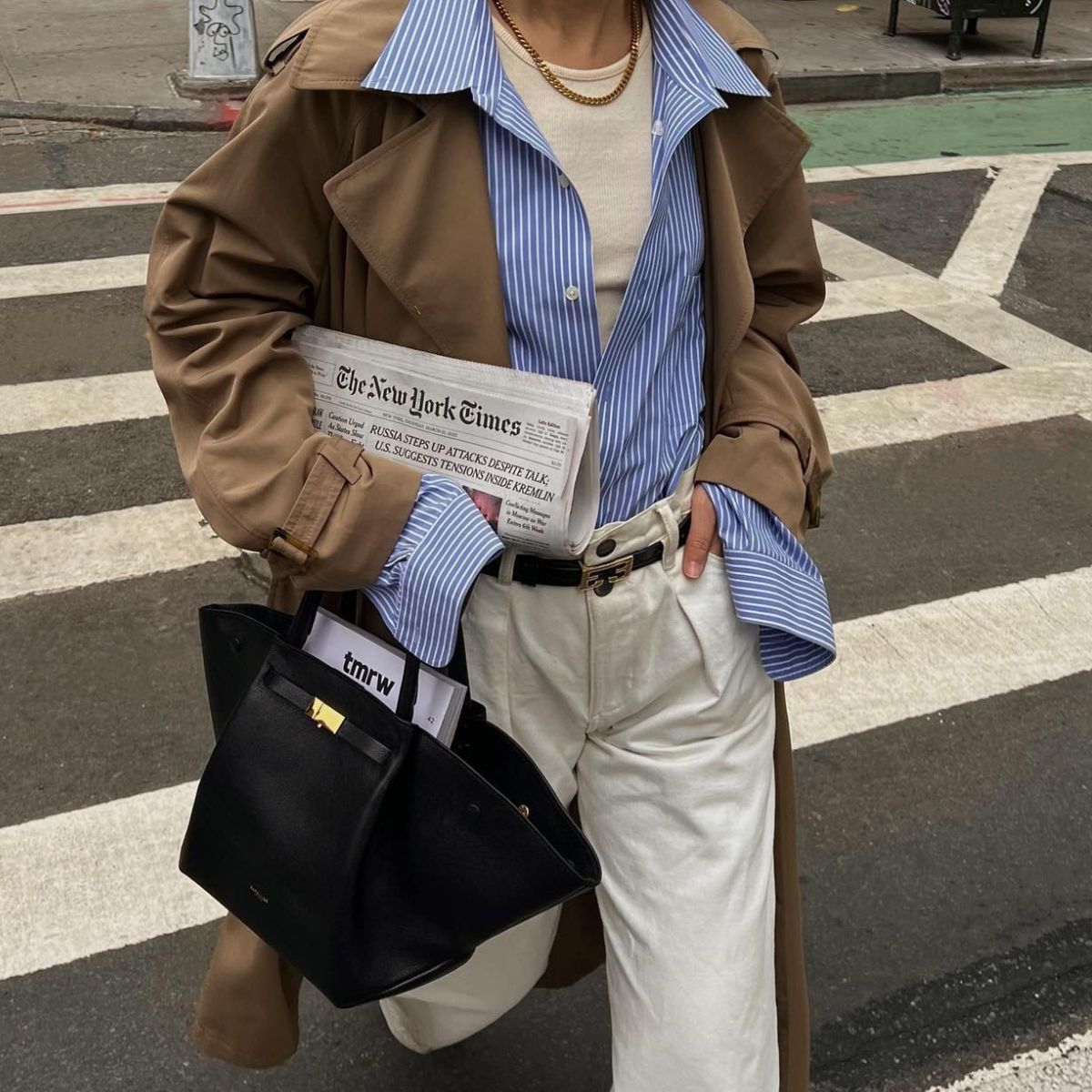 Woman wears beige trench coat, striped shirt, white jeans and carries black bag