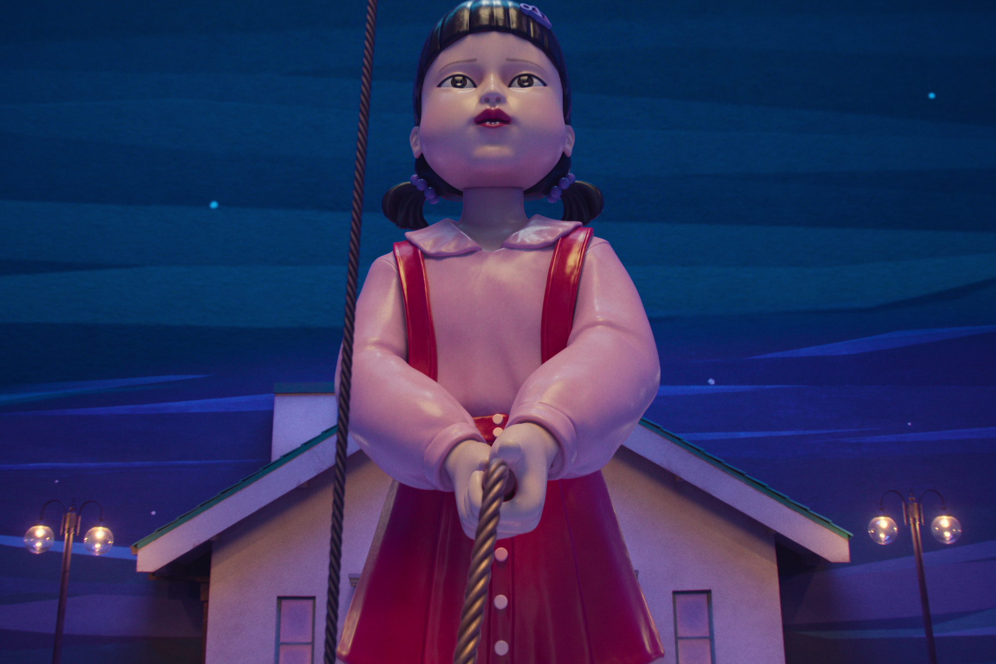Young-hee, the giant doll, holds a rope while standing in front of a house, street lamps, and a painted sky, in Squid Game S3.