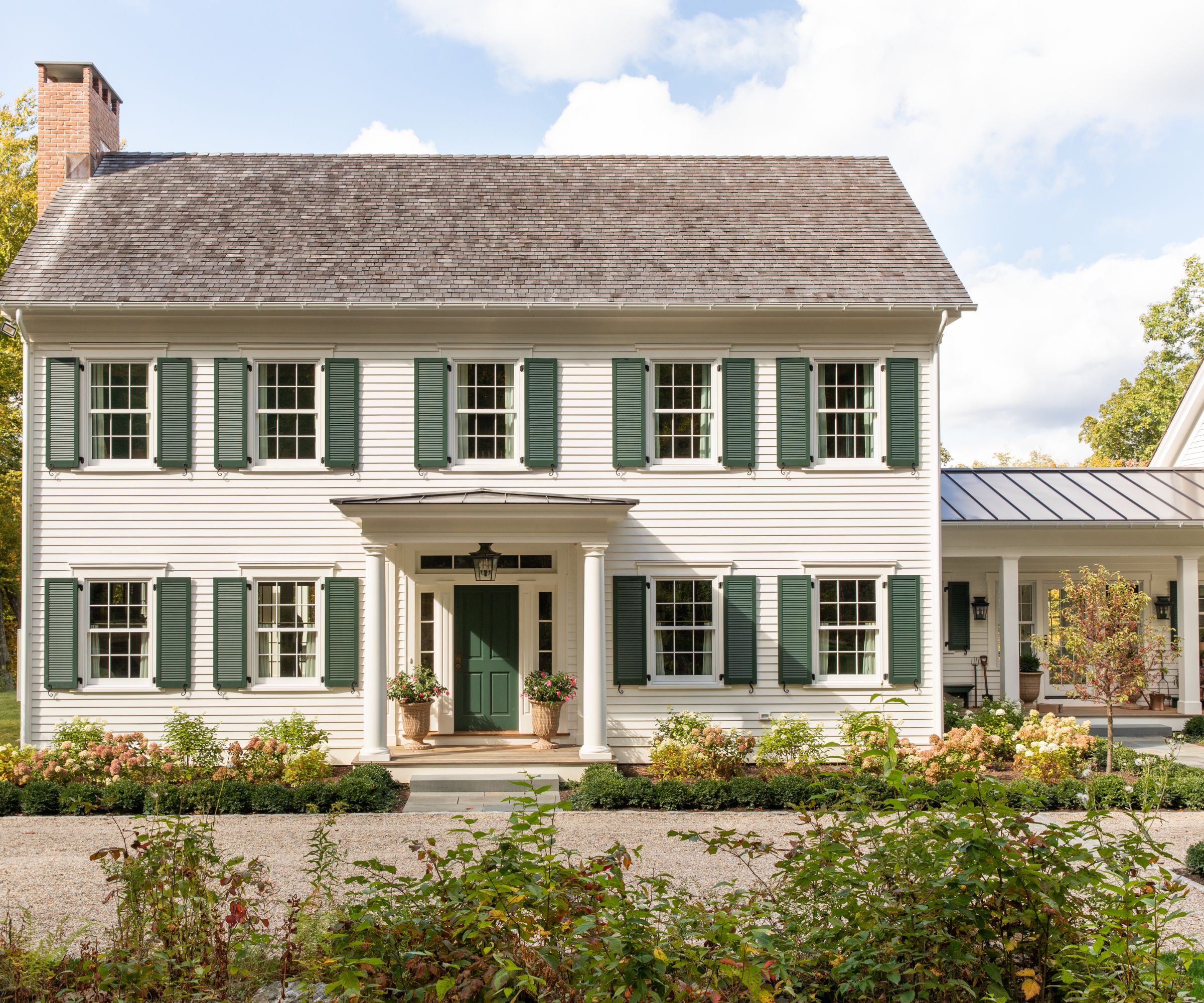 Colonial-style house with white painted exterior, green shutters, a pillared entrance with green front door and potted plants, shrubbery around the property and a side extension.
