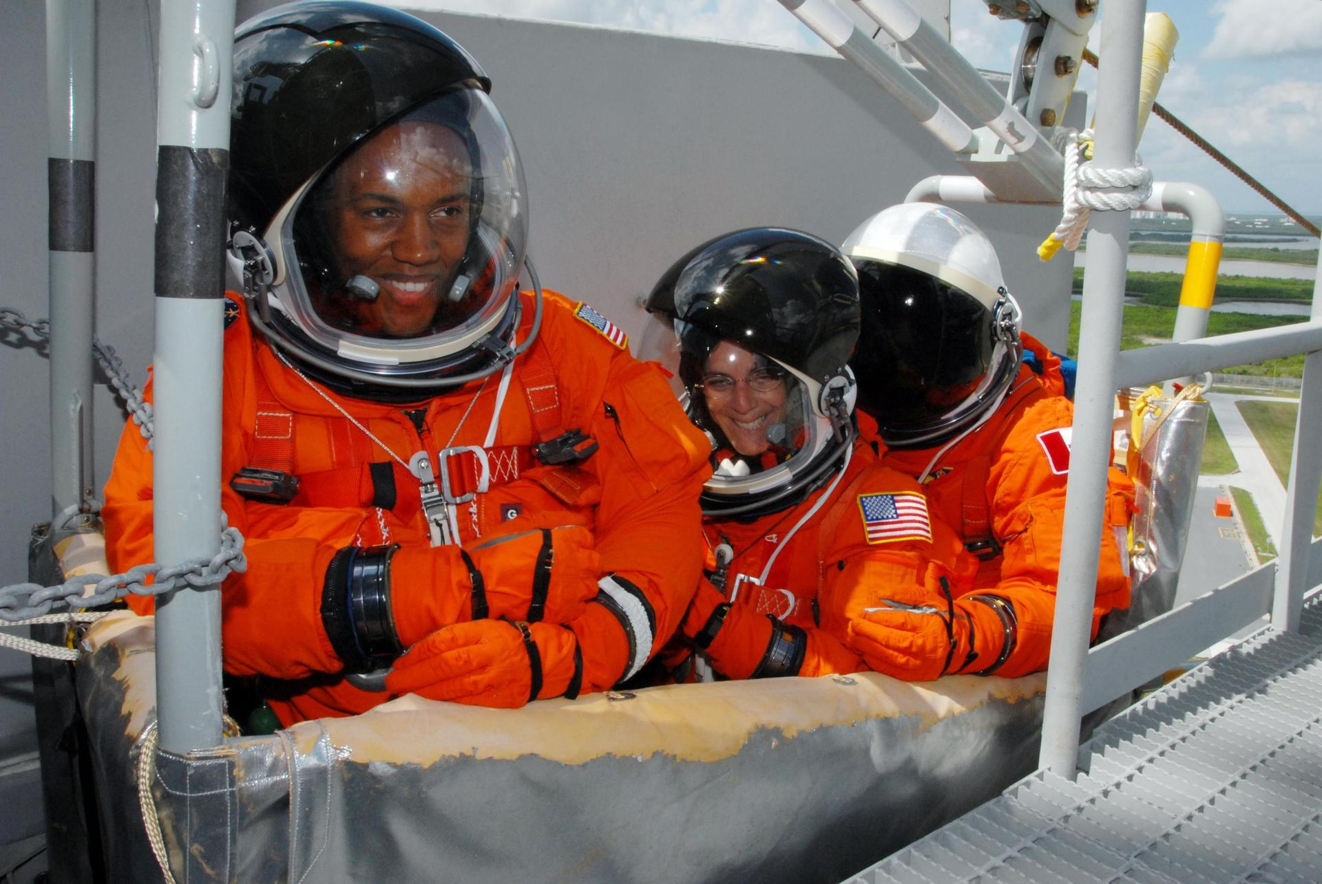 three people stand in a gurney wearing bright orange spacesuits.