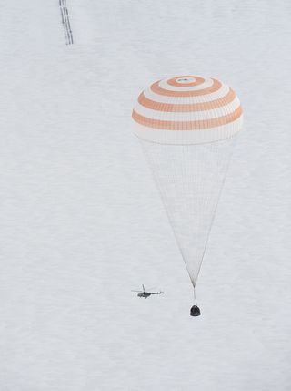 As the Soyuz TMA-16 spacecraft returns to Earth, a recovery helicopter circles the spacecraft on a snowy, windy day.