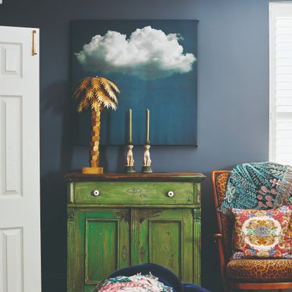 A dark blue-painted living room with a green vintage cabinet, a gold palm tree-shaped table lamp and a painting of a cloud