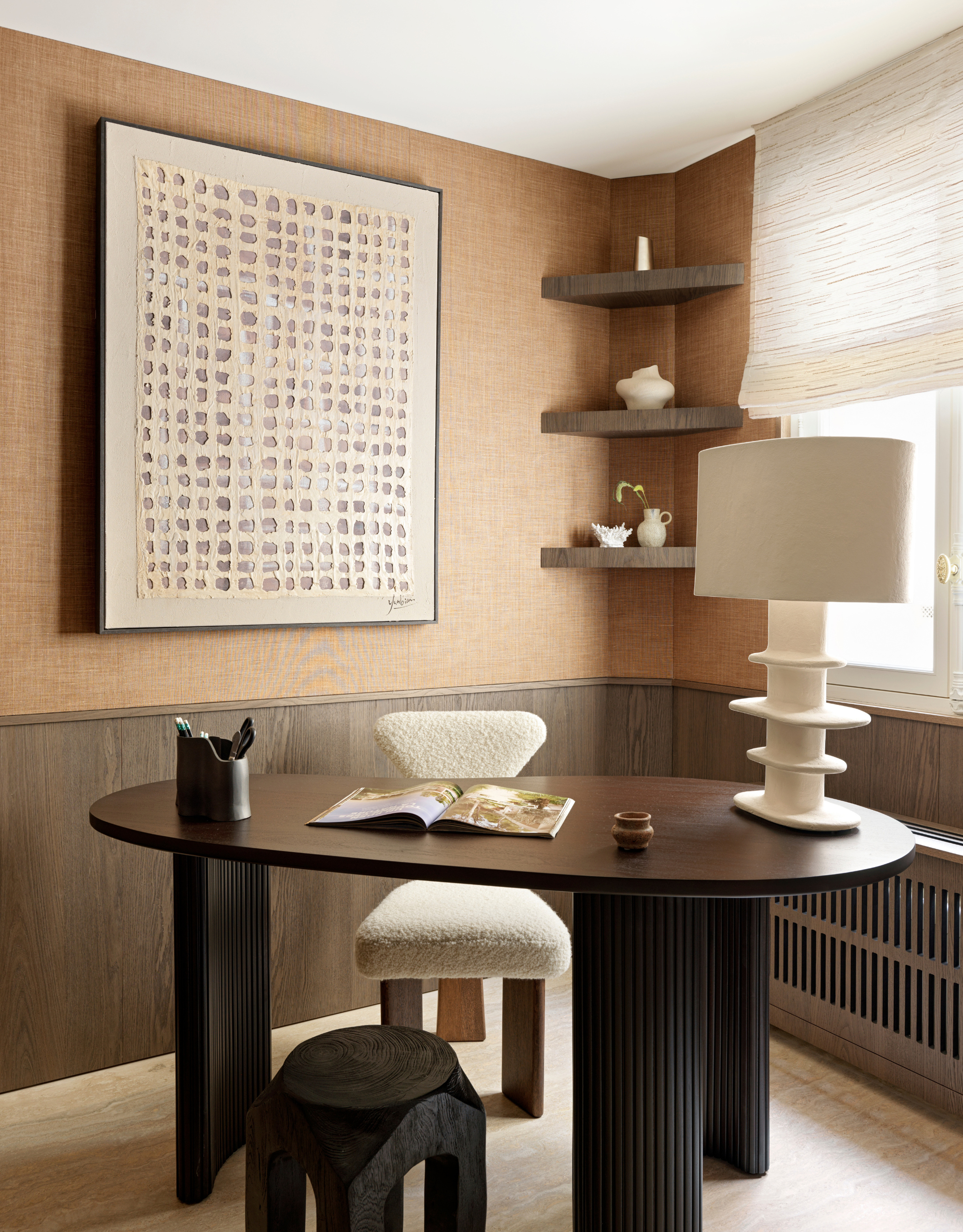 A brown home office with a bean-shaped desk with ribbed legs, a wooden stool, and a boucl&amp;eacute; seat. Also pictured is a large white table lamp, a wooden pen holder, and an open book.