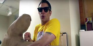 Johnny Knoxville with sunglasses on and a yellow shirt holding a giant hand and getting ready to hit the Jackass guys with it.