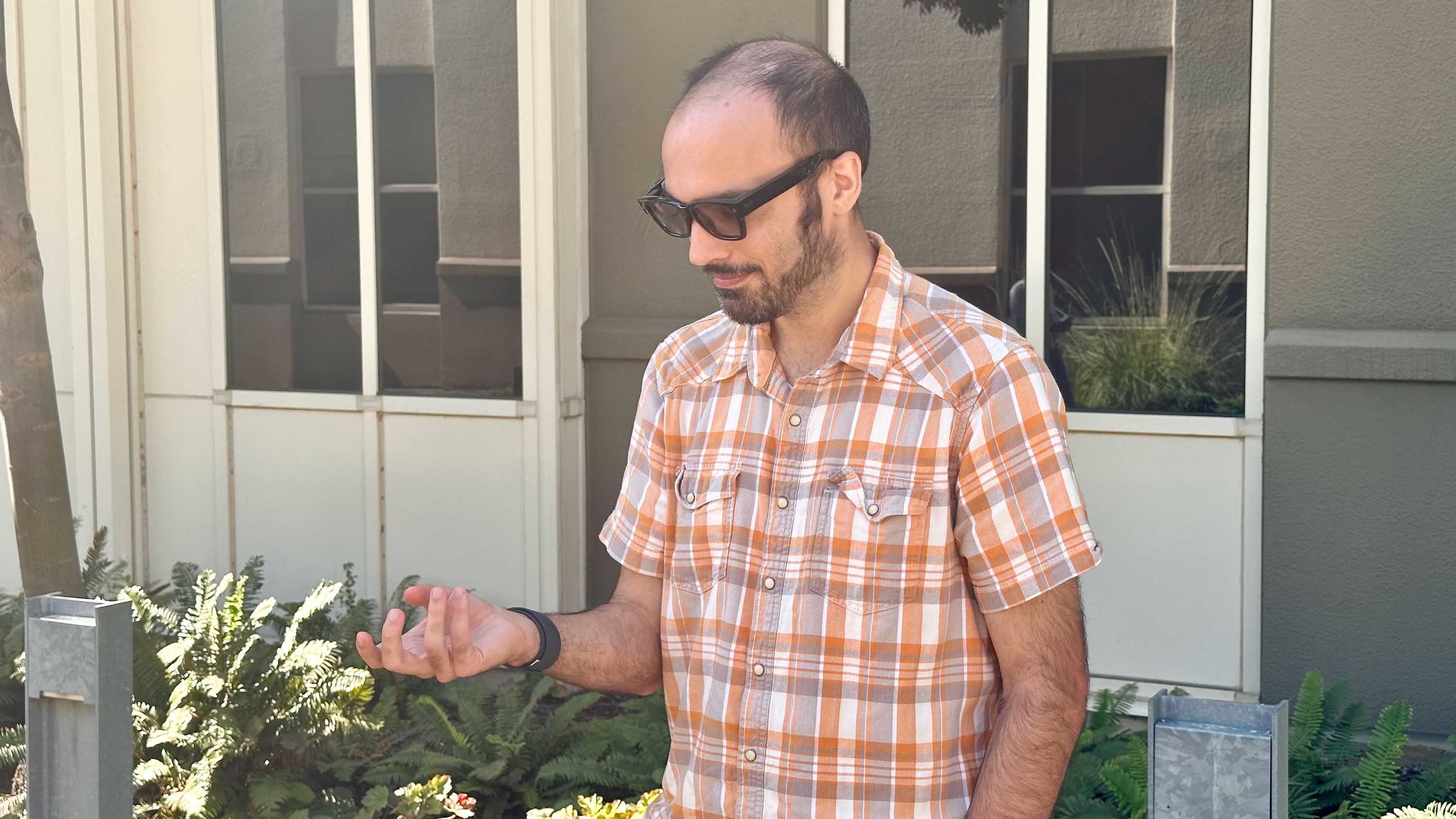 A photo of a man wearing the Meta Ray-Ban Display glasses, looking down at his hand while making a gesture tracked by the sEMG band.