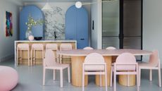 colorful kitchen with curved blue cabinets, marble backsplash and countertop with timber kitchen island, pink bar stool, gray floor, with timber and stone dining table with pink dining chairs and fluted glass doors leading to the hallway