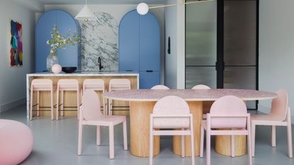 colorful kitchen with curved blue cabinets, marble backsplash and countertop with timber kitchen island, pink bar stool, gray floor, with timber and stone dining table with pink dining chairs and fluted glass doors leading to the hallway