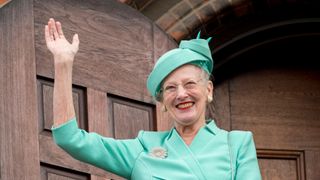 Queen Margrethe II of Denmark waves from a balcony in a blue suit and hat.