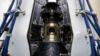 A large metal cylinder covered with black plastic is stood between blue scaffolding.