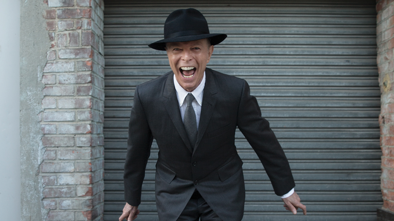 A photograph of David Bowie standing outside a metal shutter smiling in a hat, 2015