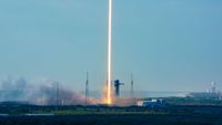A time lapse photo of a daytime rocket launch shows the rocket's fiery plume as a single orange and white beam heading skyward off the launch pad into the blue sky.