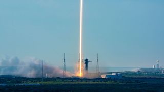 A time lapse photo of a daytime rocket launch shows the rocket's fiery plume as a single orange and white beam heading skyward off the launch pad into the blue sky.