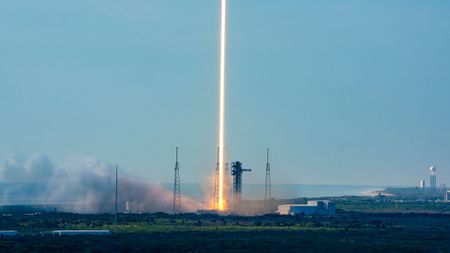 A time lapse photo of a daytime rocket launch shows the rocket's fiery plume as a single orange and white beam heading skyward off the launch pad into the blue sky.