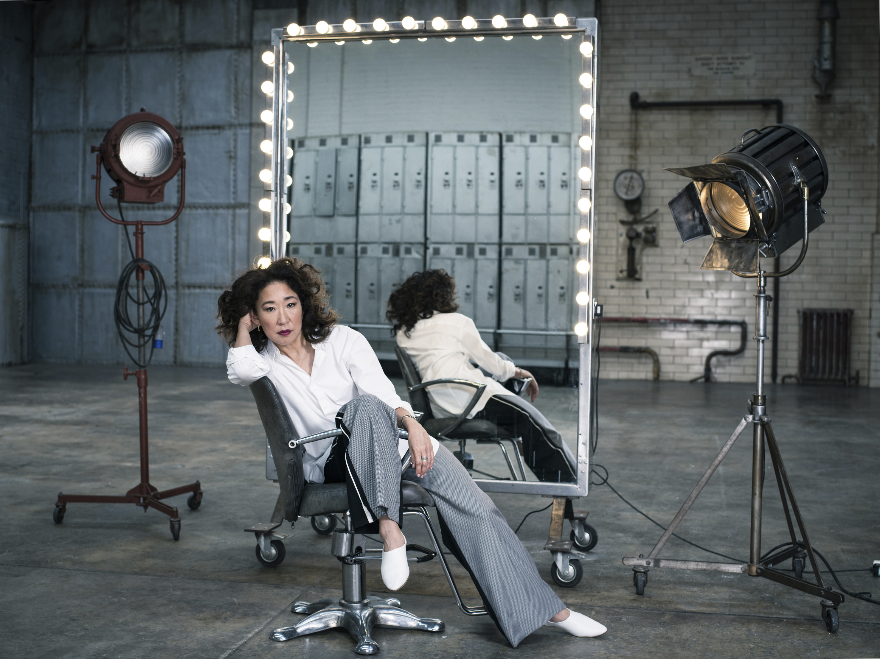 Sandra Oh sits in a barber's chair in a large industrial space, her reflection visible in a bulb-framed mirror behind her, flanked by two vintage studio lights on stands.