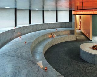 Interior view of a hamman at The Standard Spa, Miami featuring curved marble seating, candles, rolled white towels, grey pebble-like flooring and a round grey table with a bowl of fruit on top
