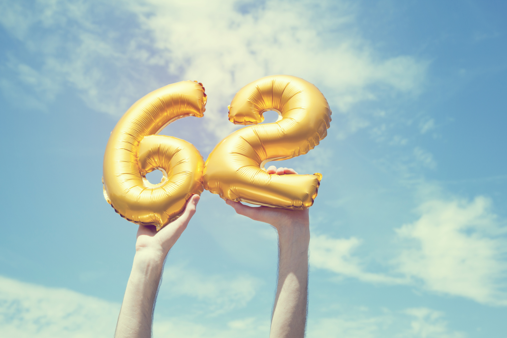 A gold foil number 62 balloon is held high in the air. The image has been taken outdoors on a bright sunny day, the sky is blue with some clouds. A vintage style effects has been added to the image.